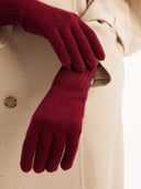 A pair of red cashmere gloves worn on a person's hands, with a beige coat in the background. #color_bordeaux