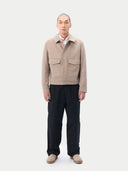 A man wearing a light beige cashmere utility jacket with a collar, front button closure, and two large flap chest pockets, layered over a white collared shirt and dark trousers, against a plain light gray studio background. #color_taupe