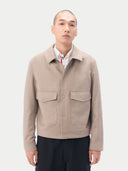 A man wearing a light beige cashmere utility jacket with a collar, front button closure, and two large flap chest pockets, layered over a white collared shirt and dark trousers, against a plain light gray studio background. #color_taupe