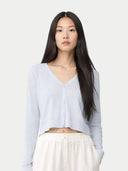 A woman with long dark hair wearing a light blue cropped V-neck cardigan and white pants, photographed against a plain light gray studio background.#color_soft-ice #ai-image