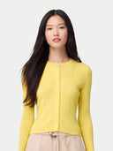 A woman with long dark hair wearing a bright yellow ribbed knit cardigan with a single button closure, paired with beige pants, against a light gray studio background. color_vivid-yellow #ai-image